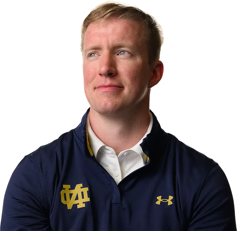 A man wearing a navy blue polo shirt with the gold 'ND' logo, representing the University of Notre Dame, looks off to the side with a thoughtful expression. He wears a white collared shirt underneath the polo shirt.